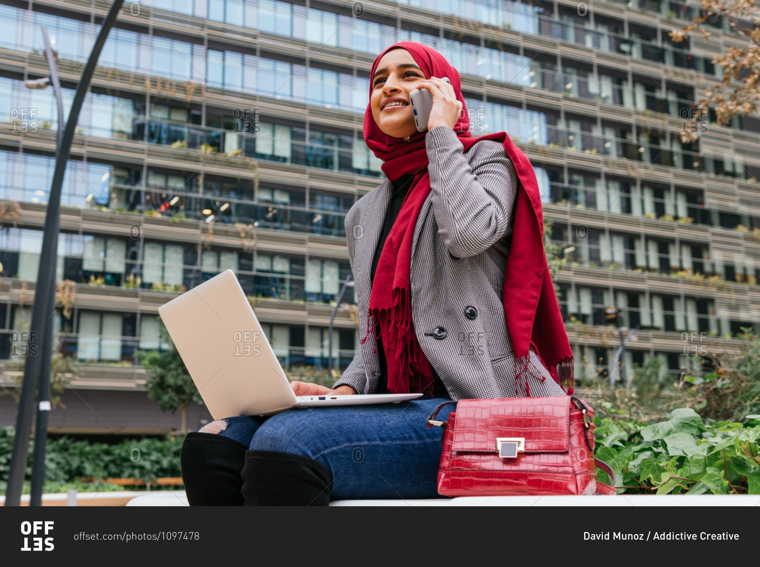 Cheerful Arab female freelancer in red hijab sitting with laptop on bench in city speaking on smartphone while working on startup project remotely