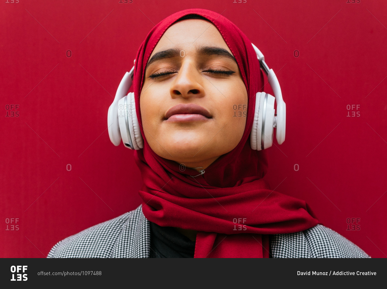 Dreamy Arab female in wireless headphones and traditional hijab standing near urban red building and enjoying music with closed eyes