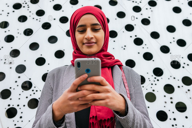 Side view of cheerful Arab female in hijab standing sending message on mobile phone while smiling and communication on social media