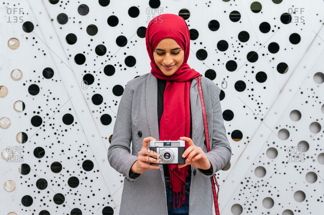 Arab female in red headscarf standing in city with retro photo camera and taking picture