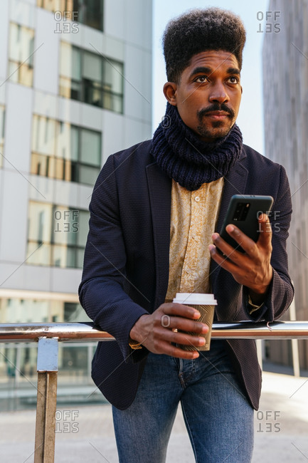 From below focused African American male entrepreneur standing in street with coffee to go and browsing smartphone while reading working emails