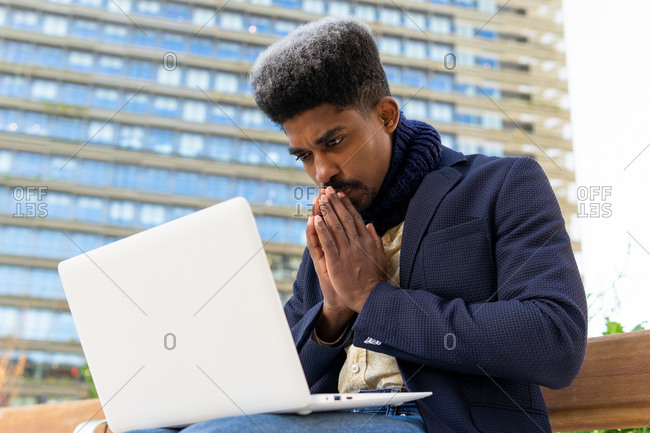 Low angle of African American male entrepreneur sitting on street with clenched fist and closed eyes while working on laptop and waiting for approval of project