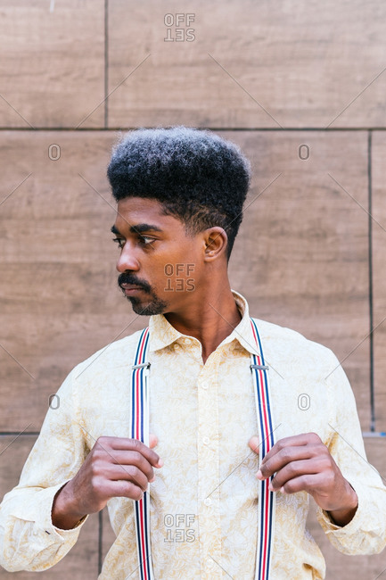 Tranquil serious stylish African American male with curly hair wearing suspenders standing on concrete wall of building and looking away while dreaming