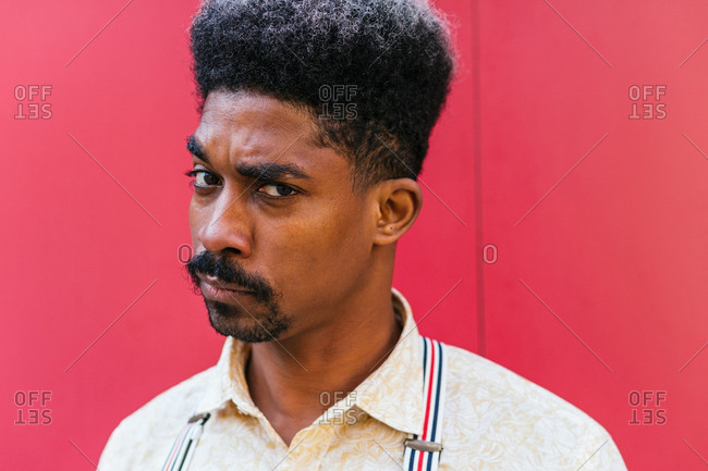 Tranquil doubtfully African American male with curly hair standing on red wall of building and looking at camera
