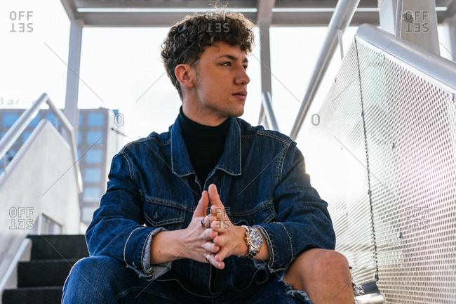 Determined young male model in denim outfit sitting on staircase in street and looking away