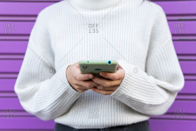 Cropped unrecognizable female millennial standing near purple wall and browsing on smartphone