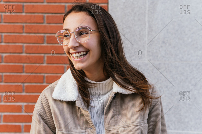 Delighted young female in glasses and trendy outfit standing on city street and looking away