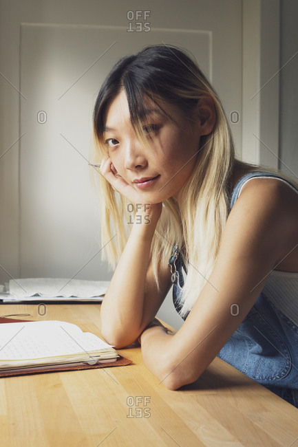 Side view of pensive Asian female sitting at table with diary and leaning on hand while looking at camera