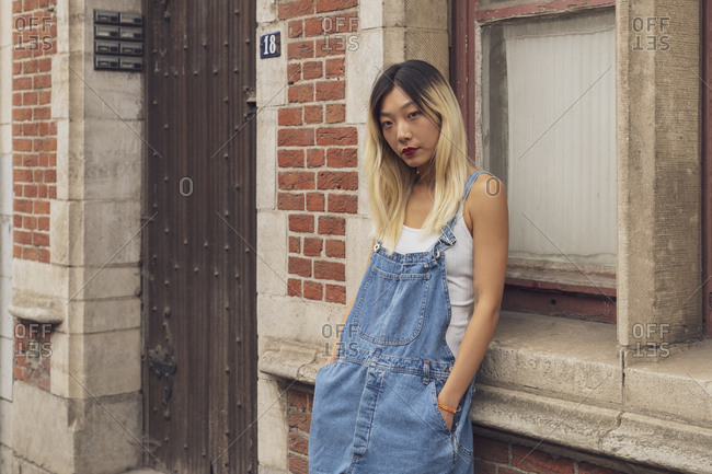 Confident Asian female wearing trendy denim overall standing with hands in pockets in city and looking at camera