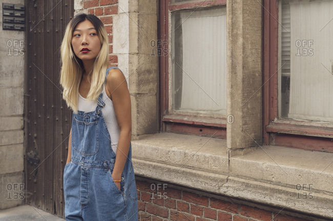 Confident Asian female wearing trendy denim overall standing with hands in pockets in city and looking away