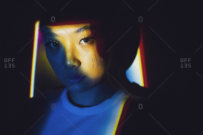 Young Asian female model with blond dyed hair and bright makeup looking at camera while standing in dark room illuminated by colorful neon lights