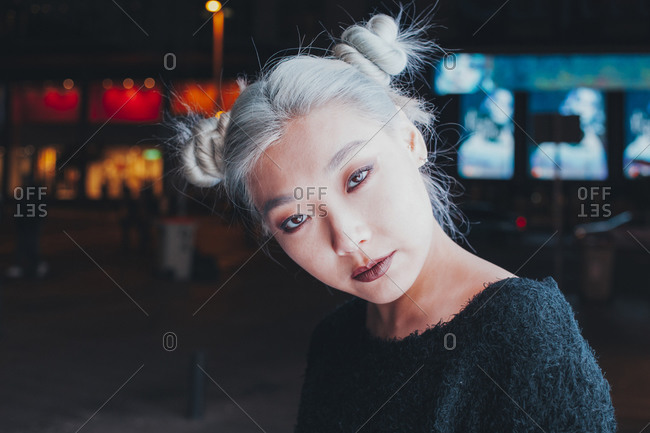 Informal Asian female adolescent with dyed blond hair and bright makeup looking at camera while standing on night city street