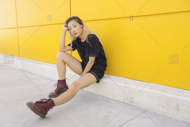 Full body side view of Asian teen female hipster in casual wear and boots sitting near yellow wall on city street
