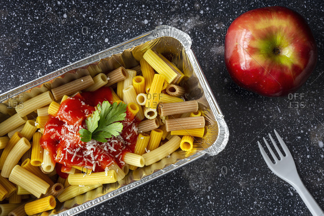 From above of delicious macaroni with ketchup and cheese placed in container for takeaway food on table with plastic fork and apple