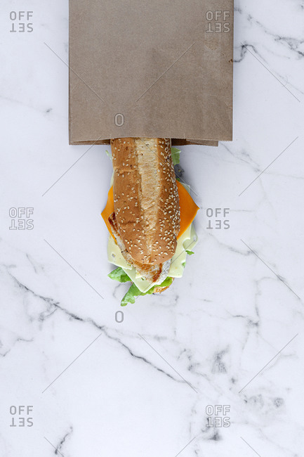 From above of yummy fresh sandwich with cheese and vegetables placed on table in takeaway paper bag