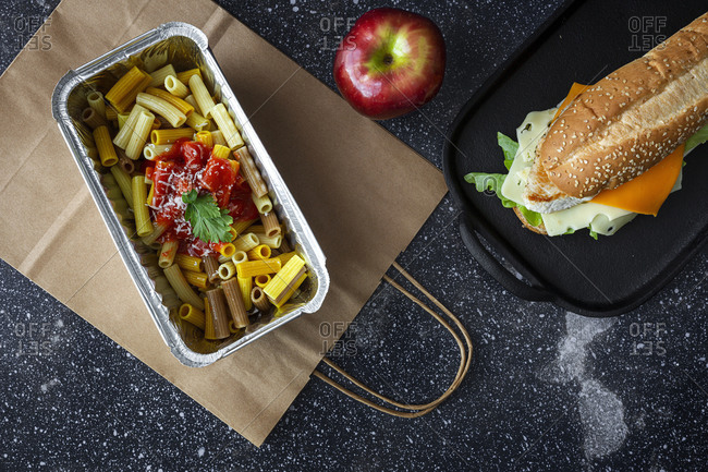 Top view composition with containers with pasta and sandwich placed with fresh red apple on table with paper bag