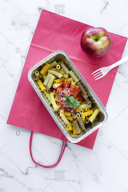 Top view of foil box with delicious pasta with tomato sauce placed near fresh red apple on table with disposable forks and paper bags representing healthy takeaway food concept