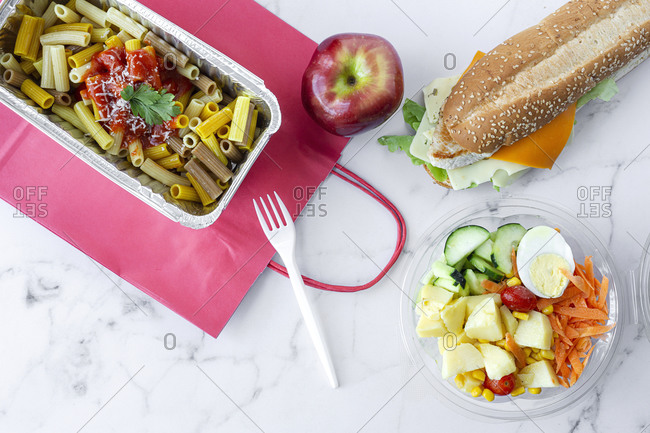 Top view composition with delicious pasta in foil box placed near salad bowl, fresh apple and sandwich with cheese and vegetables on table with paper bag prepared for takeaway lunch
