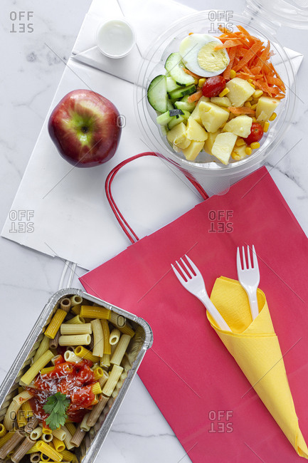 Top view composition with delicious pasta in foil box placed near salad bowl and fresh apple on table with paper bag prepared for takeaway lunch