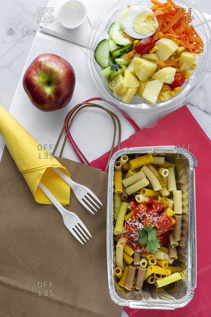 Top view composition with delicious pasta in foil box placed near salad bowl and fresh apple on table with paper bag prepared for takeaway lunch