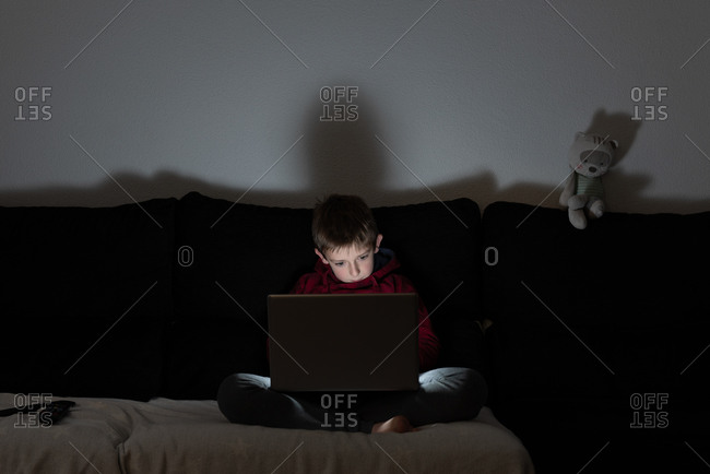 Pensive preteen boy in casual wear sitting on sofa and browsing laptop during free time at home in the evening