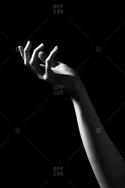 Black and white of crop anonymous female with slim arm on dark background in studio reaching towards light