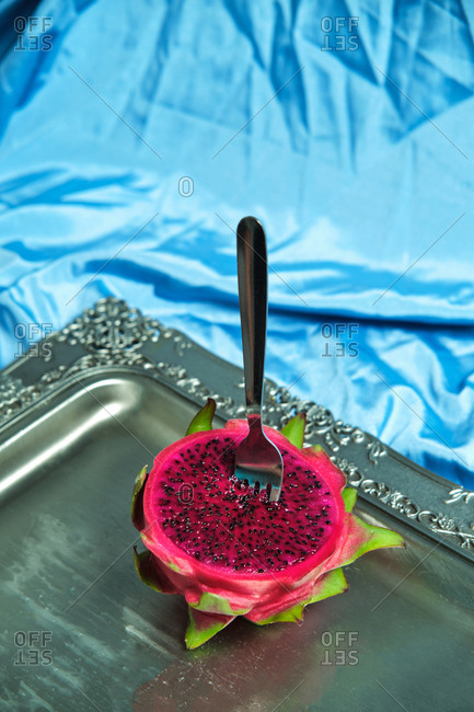 High angle of half of fresh juicy dragon fruit with fork placed on metal tray on blue wrinkled textile in studio