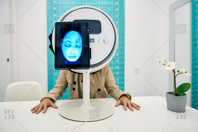 Unrecognizable woman sitting at table in facial skin diagnostics equipment with tablet during procedure in modern beauty clinic