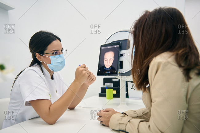 Side view of cosmetologist and customer sitting at table with facial skin diagnostics equipment while looking at tablet and discussing results of analysis in beauty clinic