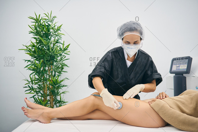 Professional masseuse doing anti cellulite massage on legs of female client while using special machine in modern beauty clinic