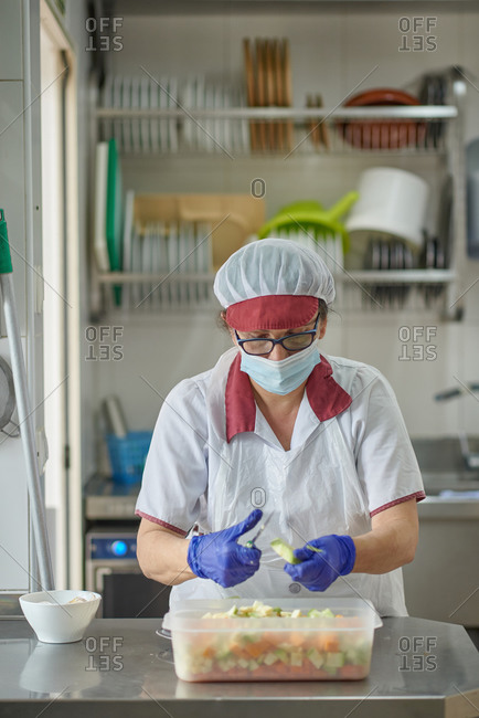 Female cook in white uniform and protective mask and gloves cutting fresh vegetables while preparing food in hospital kitchen during coronavirus pandemic
