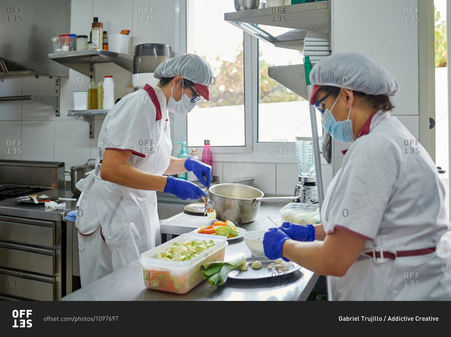 Side view of unrecognizable female cooks in protective masks and gloves preparing meal for patients while working in hospital kitchen during coronavirus pandemic