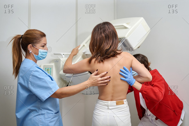 Female medics in masks and uniform preparing woman with naked torso for chest radiology while standing in medical room in clinic
