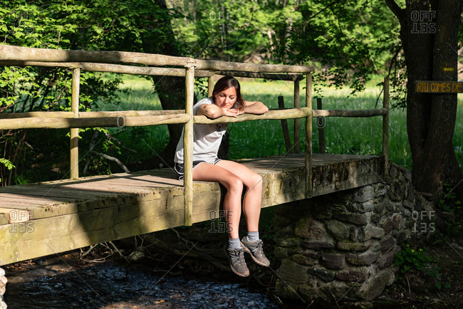 Calm female sitting on old bridge over river in woods and enjoying nature in summer
