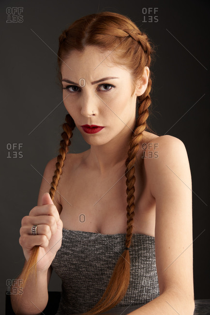 Stylish female model with red hair and braids sitting in studio on dark background and looking at camera