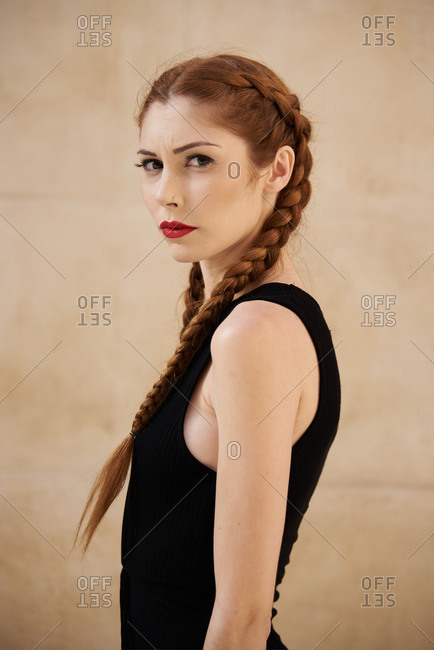 Side view of stylish female model with red hair and braids sitting in studio on dark background and looking at camera