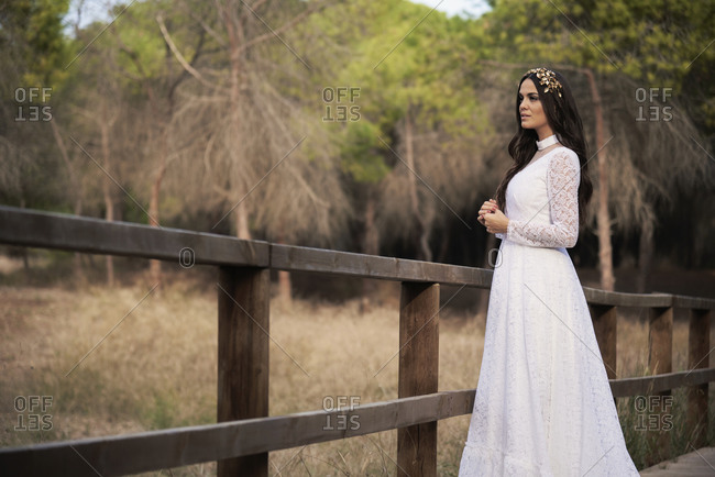 Side view of happy elegant young female in boho bridal maxi dress and wreath standing on wooden walkway in green woods and looking away