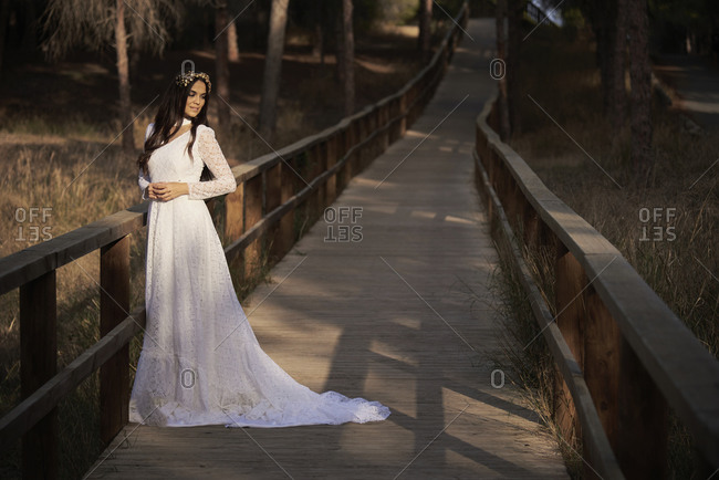 Full body side view of happy elegant young female in boho bridal maxi dress and wreath standing on wooden walkway in green woods and looking away