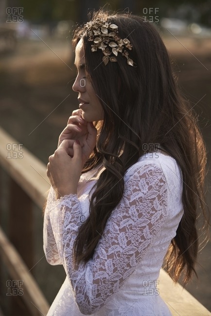 Side view of elegant young female in boho bridal maxi dress and wreath in green woods and looking away