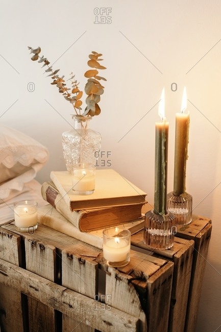 Fragment of interior of bedroom with rustic wooden handmade bedside table with dry plants in glass vase placed on stack of old books near burning candles