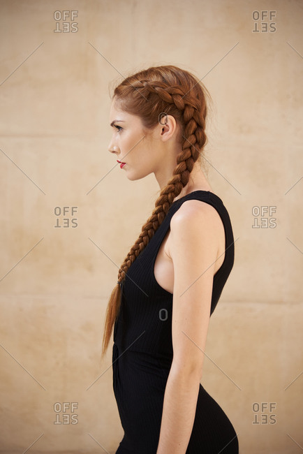 Side view of stylish female model with red hair and braids sitting in studio on dark background and looking away