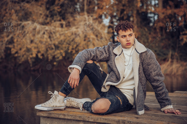 Young man wearing jacket sitting on jetty against lake at autumn forest