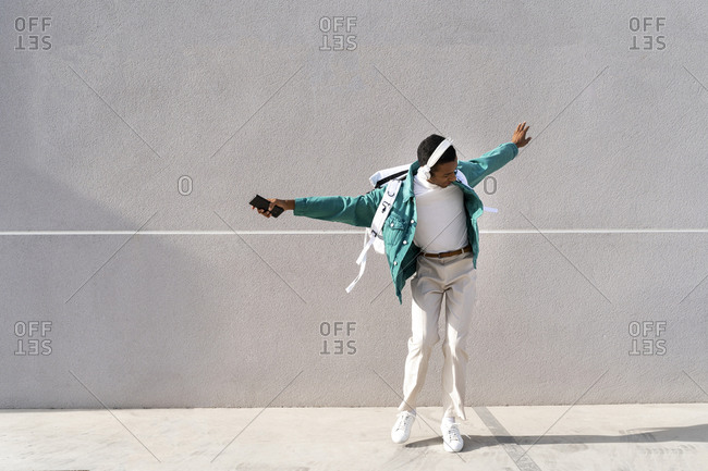 Carefree man with headphones and mobile phone dancing with arms outstretched against gray wall