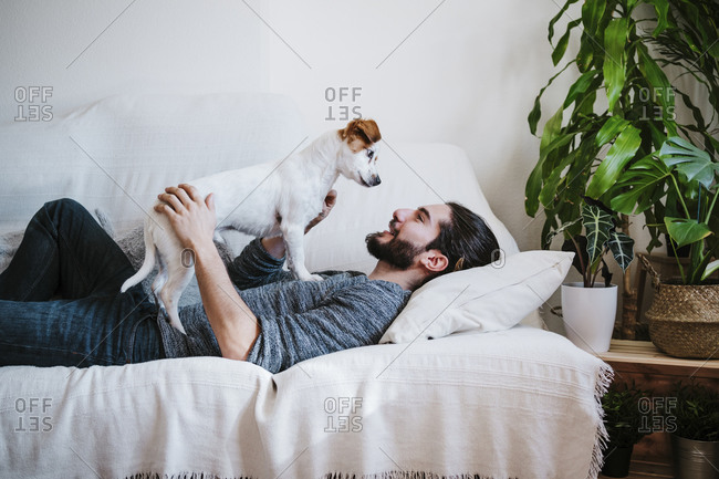 Smiling man playing with dog while lying on sofa at home