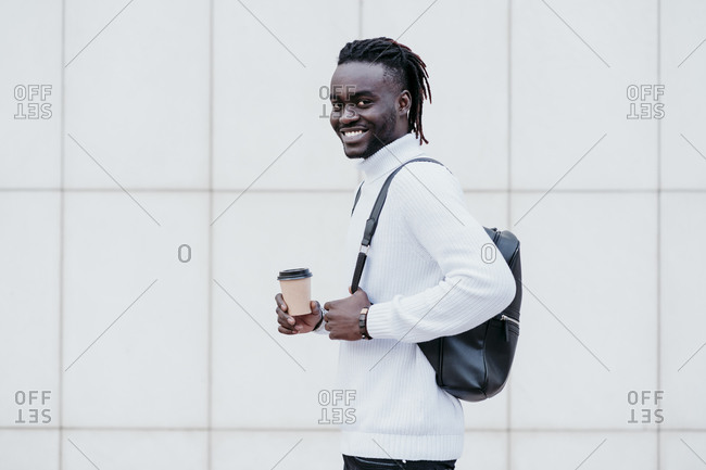 Young businessman carrying backpack while holding disposable coffee cup against white wall