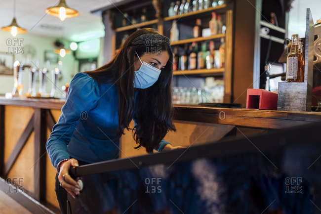 Mid adult woman wearing protective face mask playing pinball in bar during pandemic