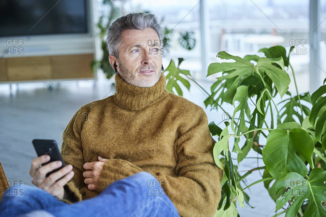 Mature man wearing in-ear headphones using mobile phone while sitting at home