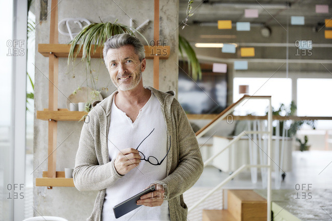 Smiling mature man holding digital tablet and eyes glasses while standing at office
