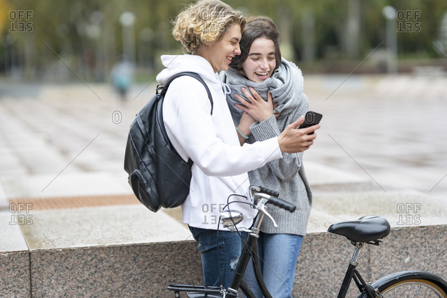 Young friends with bicycle laughing while watching video through smart phone on footpath