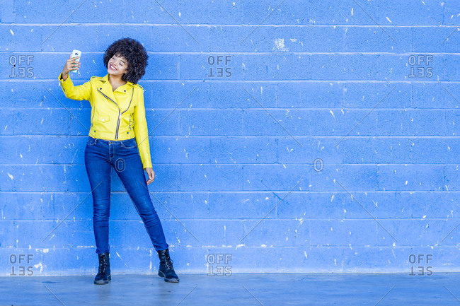 Happy fashionable woman in yellow leather jacket taking selfie against blue wall